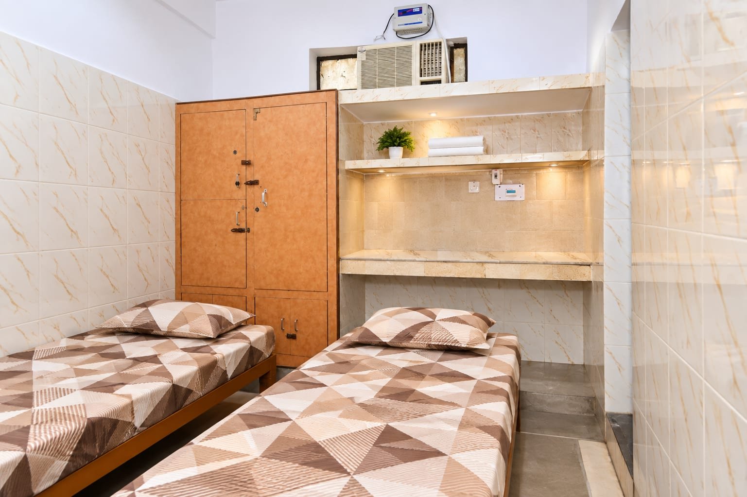 A double-sharing room with an air conditioner above the study ledge, a wooden almirah and two single beds
