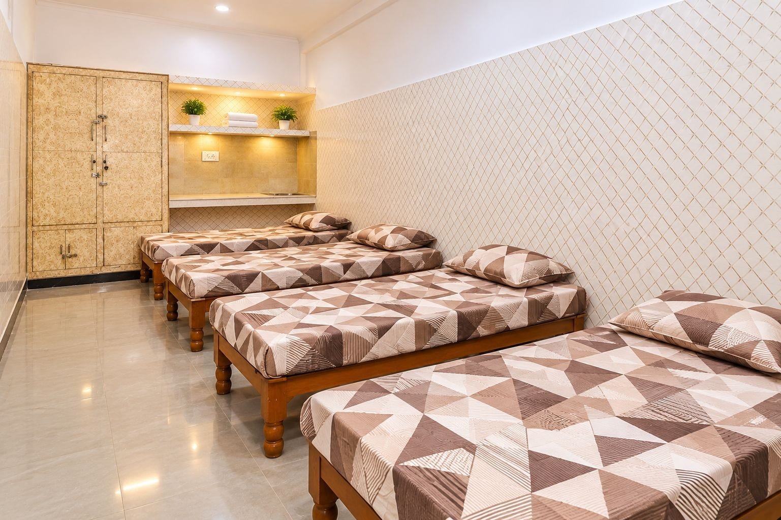 A four-sharing room with four single beds, patterned bed linen, a wooden almirah and a study counter along the wall